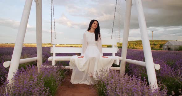 a Gentle Girl in a White Light Dress Swings on a Swing in a Field with Flowers alt