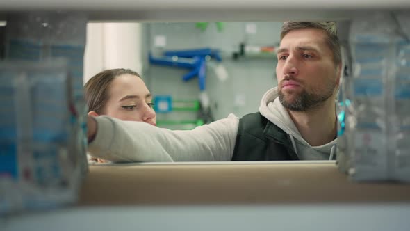 Focused Caucasian Couple Standing Behind Shelf in Hardware Store Talking and Choosing Goods alt