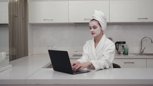 A Girl in a White Robe and Face Mask Works Behind a Laptop Sitting at Home