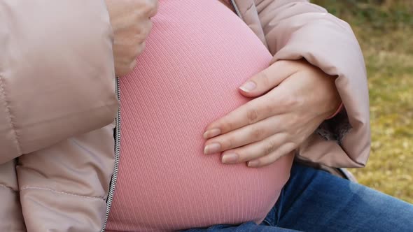 A pregnant woman sitting in the park for a walk and stroking her belly.