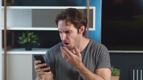 Shocked Man Reading Terrible News on His Smartphone at Home Apartment Background. Unpleasantly alt