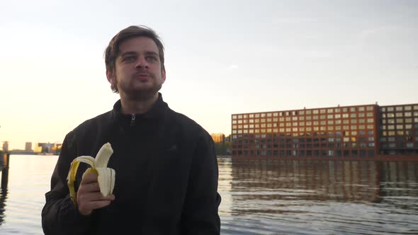 Handsome man eating a banana and smiling on a pier during sunset alt