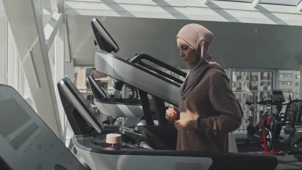 Woman in Hijab Drinking Water after Treadmill Workout in Gym alt