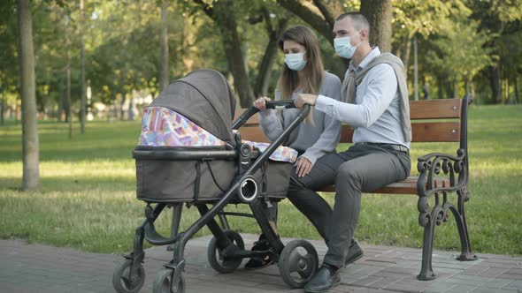 Happy Young Couple of Parents in Covid-19 Face Masks Rocking Baby Stroller and Talking. Wide Shot of alt