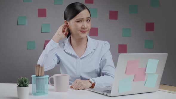 Asian woman working on a laptop itching and putting a finger into her ear sitting