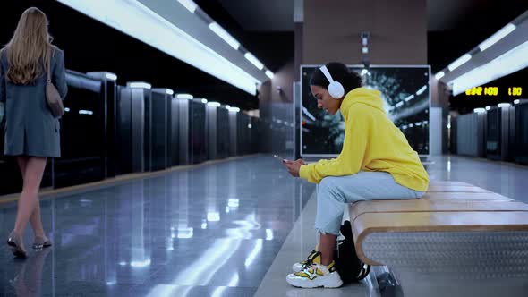 Young Girl Sitting in Modern Metro Station Waiting for Train Wearing Headphones alt