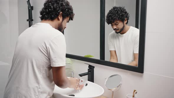 Young Indian Man Washes Hands Under Faucet in Bathroom alt
