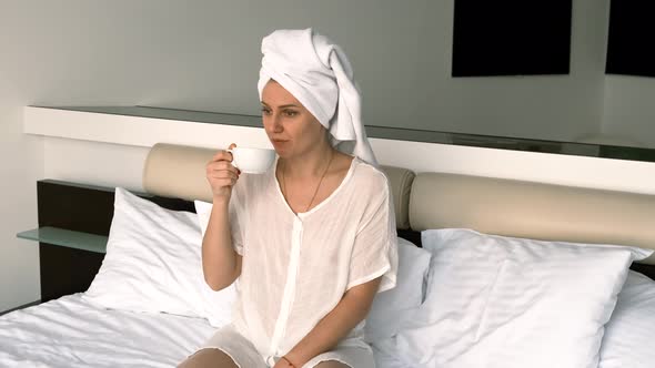 Girl Wearing Bathrobe and Towel Drinks Coffee While Sitting on the Bed