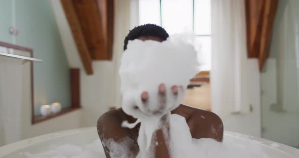 Smiling african american attractive woman taking bath and blowing out foam in bathroom alt