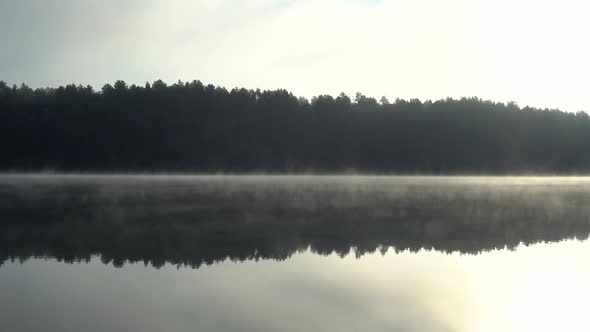 Morning Fog Blows Slowly Across a Clam Lake with Forest Reflecting in the Water