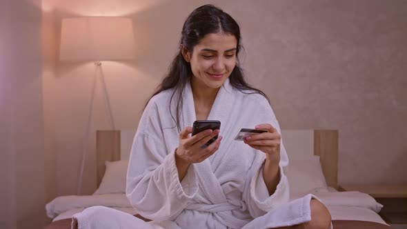 Smiling Young Woman Customer Holding Credit Card and Smartphone Sitting on Couch at Home alt