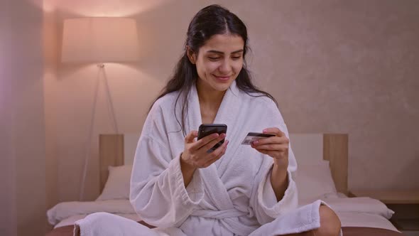 Smiling Young Woman Customer Holding Credit Card and Smartphone Sitting on Couch at Home