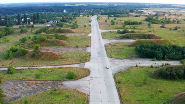 Flying over a car that is traveling on an airplane runway. Forest and grass on the sides. alt