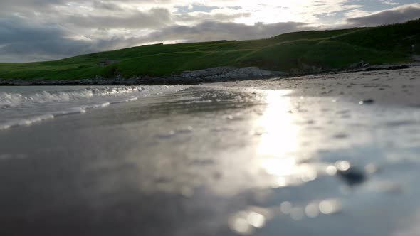 A low angle shot of waves gently crash up against a sandy beach very close to the camera as the sun alt