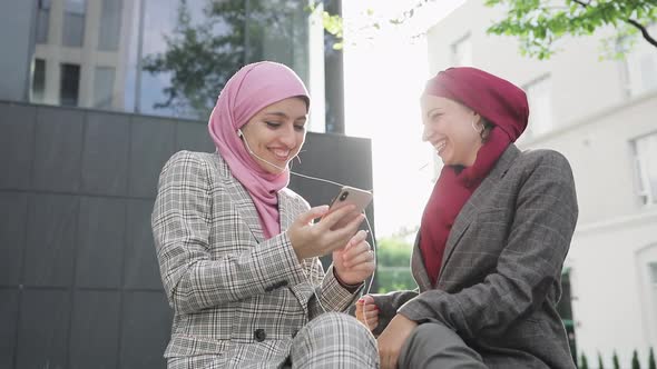 Two Young Muslim Women Wearing Hijab Headscarf Listen Music on Phone By Earphones, Sitting Together
