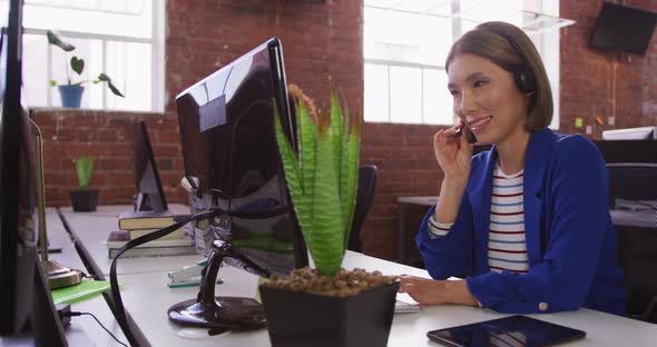 Diverse male and female business colleagues wearing headsets sitting at desks having video calls alt