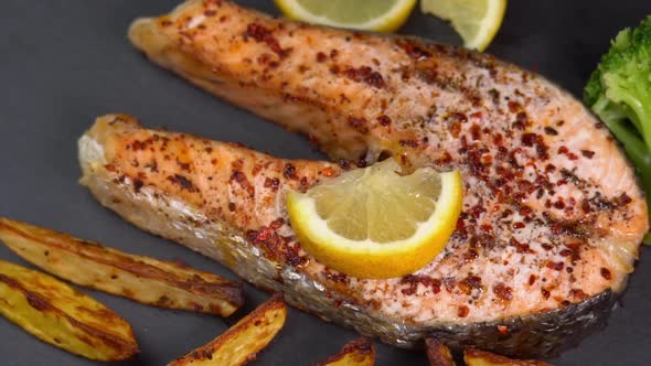 Grilled salmon fillets spinning on a black plate with lemon slice, french fries and boiled broccol alt