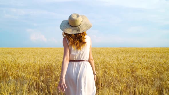 Red haired girl in sumnmer dress and straw hat walking on wheat field alt