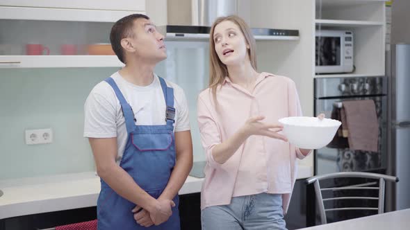 Sad Caucasian Woman and Service Man Looking at Water Drops Falling From Ceiling and Talking alt