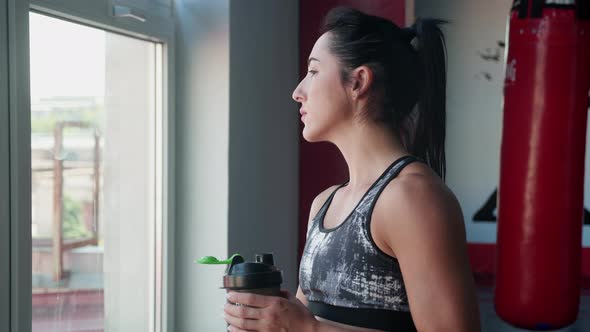Young Woman in Sport Outfit is Standing Near the Window Serfing News in Smartphone and Drinks alt