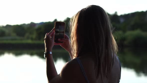 Backside shot of a girl taking photo of summer lake landscape; bokeh nature background alt