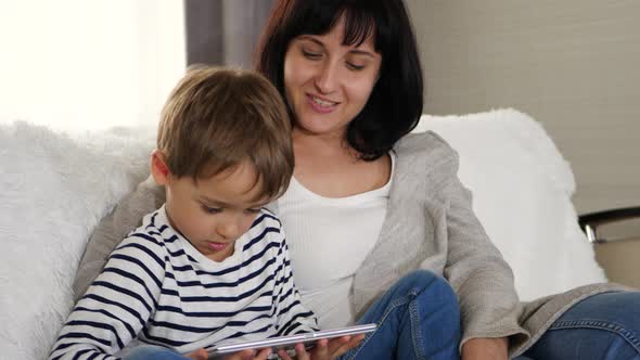Mother and Son Spend Time Together Sitting on the Sofa and Watching a Movie with a Tablet alt