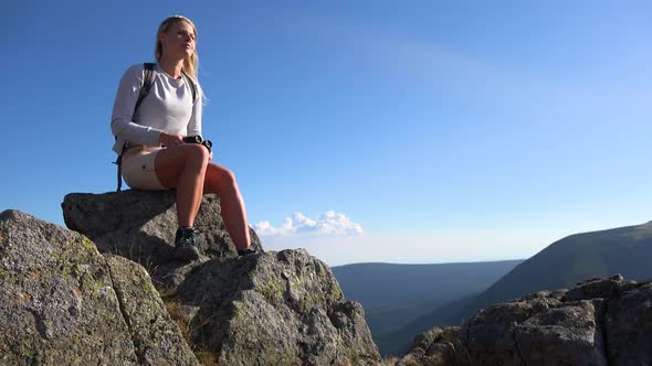 A Young Beautiful Woman Sits on a Rock on a Hilltop and Looks Around Through Binoculars alt