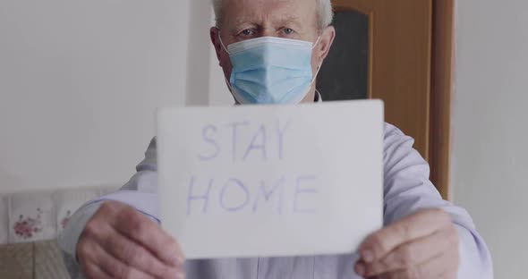 Worrid Senior Man in Mask on Isolation Shows at Camera Inscription "STAY HOME" alt