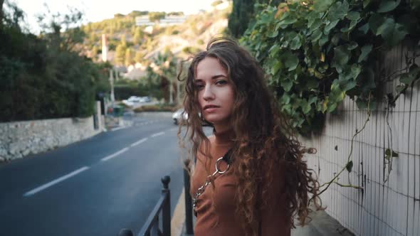 Curly Girl Looks and Enjoys the View. Hair Flies in the Wind. Sunny European Landscape. Young Woman alt