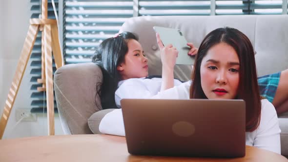 Mother is working on a computer and her daughter is playing games with her tablet alt