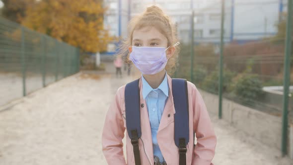 A Little Schoolgirl Girl in a Protective Medical Mask is Walking From School with a Backpack on Her