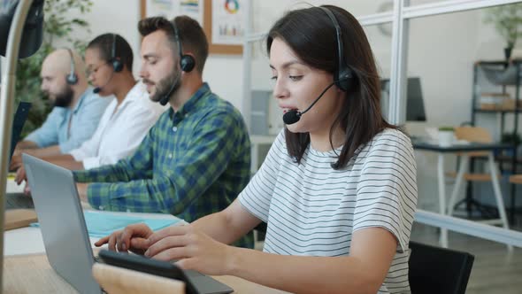 Cheerful Young Woman Call Center Worker Talking and Typing Using Modern Equipment with Colleagues in alt