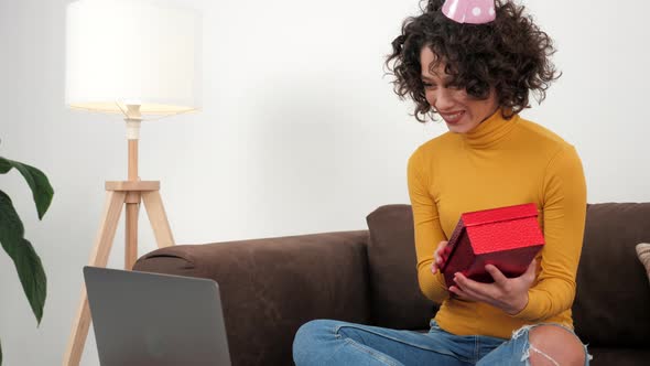 Happy Woman in Party Hat Opens Gift Box and Surprised Sitting in Couch at Home alt