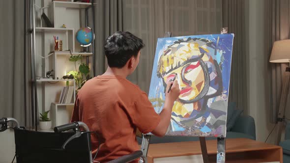 Hind View Of An Asian Artist Boy In Wheelchair Thinking And Painting A Girl's Face On The Canvas alt