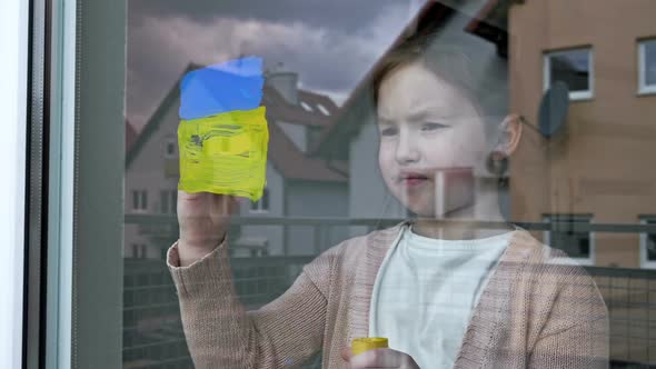 Little Girl Draws the Flag of Ukraine on the Window alt