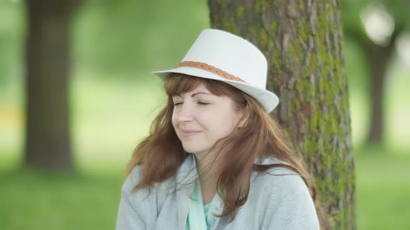 Happy Woman in a Hat Sitting Under a Tree in the Park Turns His Head, Smiling and Looking 