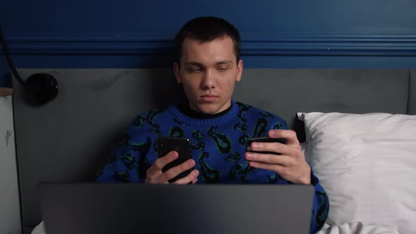 Young Caucasian Man Holding Credit Card and Smartphone Sitting on Bed at Home. Man Shopper Using alt
