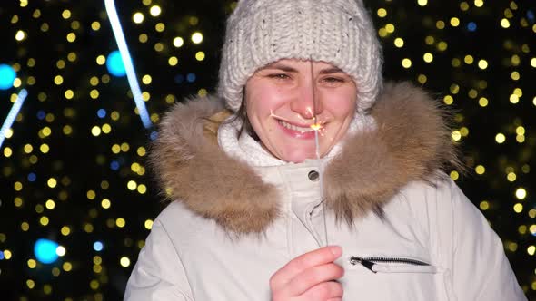 A Woman Holds a Sparkler and Smiles Standing Outside a Christmas Tree in the Open Air alt