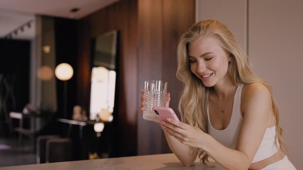 A Young Blonde in the Kitchen Drinks Water From a Glass and Chats By Phone alt
