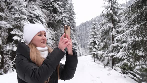 Blonde Girl Holds a Smartphone in Her Hands and Takes Pictures of a Beautiful Winter Forest alt