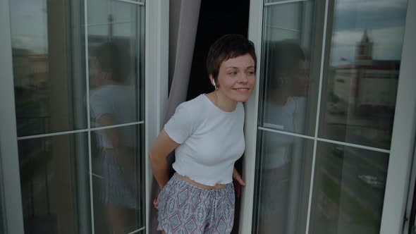 A Young Woman Listens to Music with Headphones and Dances on the Balcony on a Day Off alt