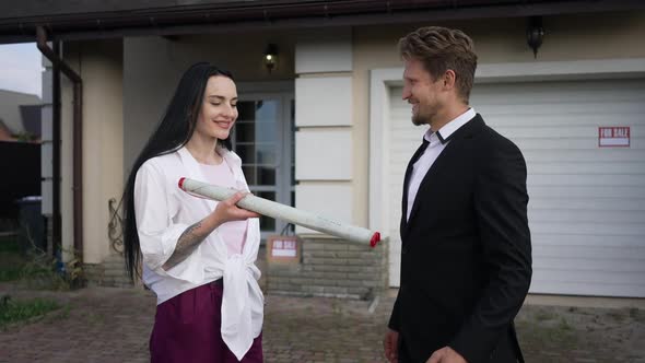 Satisfied Smiling Woman Standing with Expert Man Outdoors Passing Blueprint alt