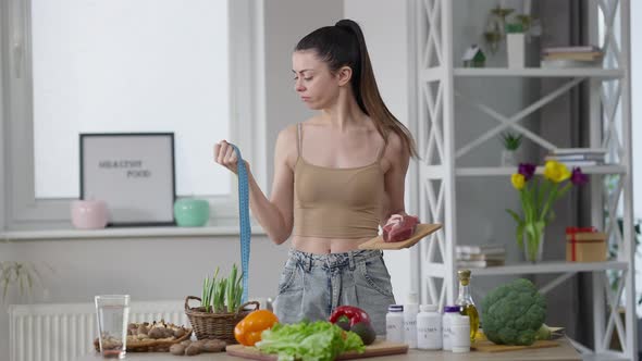 Unsure Caucasian Young Slim Woman Holding Measuring Tape and Raw Meat Making Decision and Looking at alt
