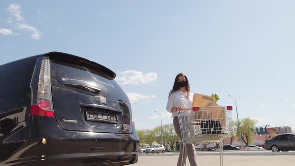 Woman in Face Mask Loading Groceries into Car alt