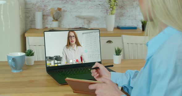 Woman Sits at Desk and Makes Notes in Notebook on Laptop Screen Positive Woman in Glasses Speaks alt