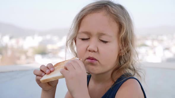 Little Blonde Girl is Eating Sandwich for Breakfast Sitting in Terrace