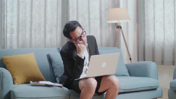 Asian Businessman In Jacket And Shorts Holding His Cheek And Sleeping While Working With A Laptop alt