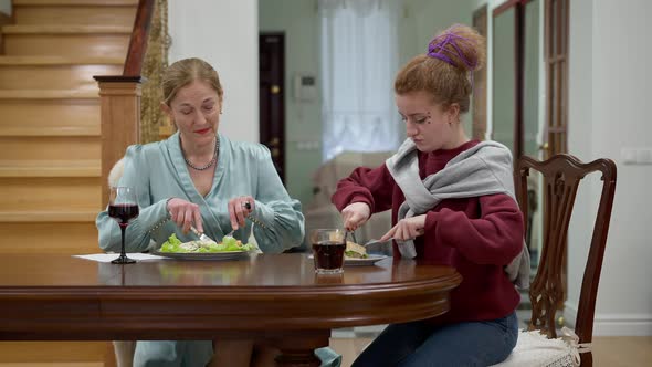 Portrait of Elegant Middle Aged Lady Teaching Hipster Woman Eating with Fork and Knife Indoors alt