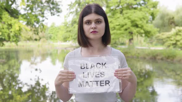 A Young Caucasian Woman Shows a Black Lives Matter Sign to the Camera in a Park alt