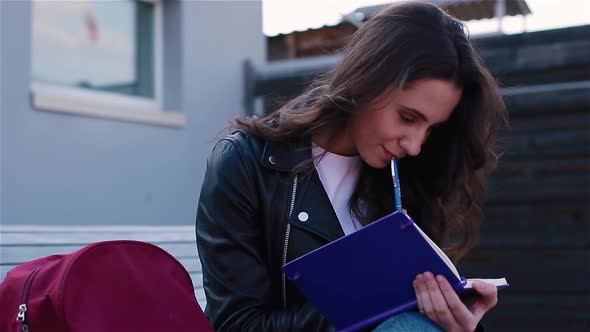 Female Student Writing in Notebook Sitting Outside on the Modern Wooden Bench alt